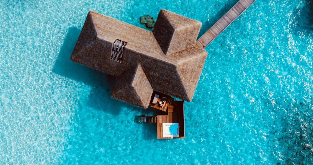 Overhead view of a thatched overwater villa with a small pool, surrounded by turquoise water.