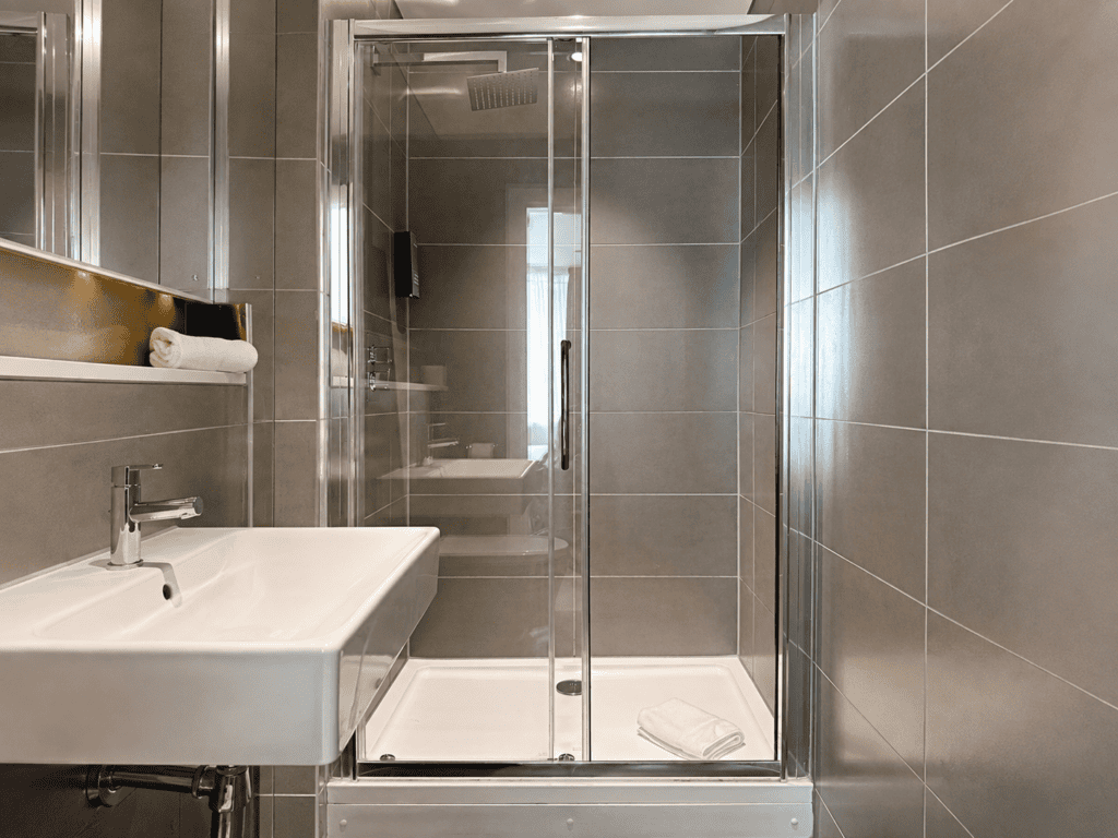 Compact en suite bathroom at Staycity Manchester Piccadilly with glass shower, wall-mounted sink and grey tiling.