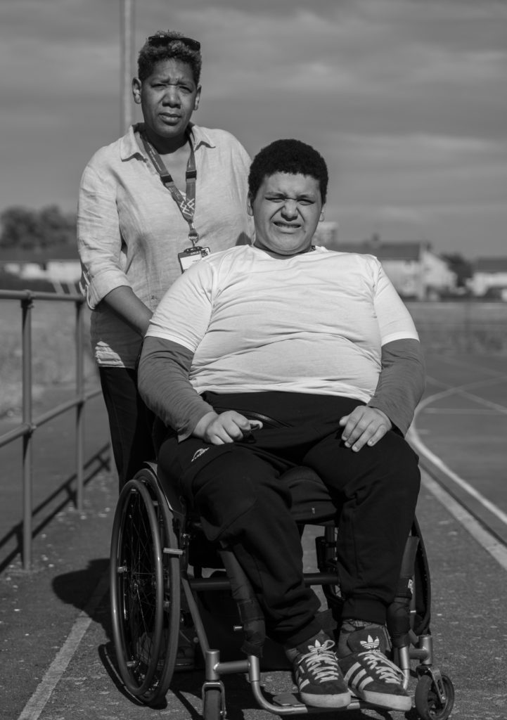 Caron McLuckie standing behind her son Emile, who is seated in his wheelchair outdoors.
