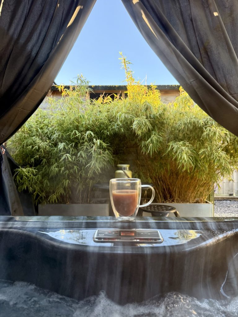 A glass mug of a hot drink on the edge of a bubbling outdoor hot tub, with dark curtains framing bamboo and a clear blue sky.