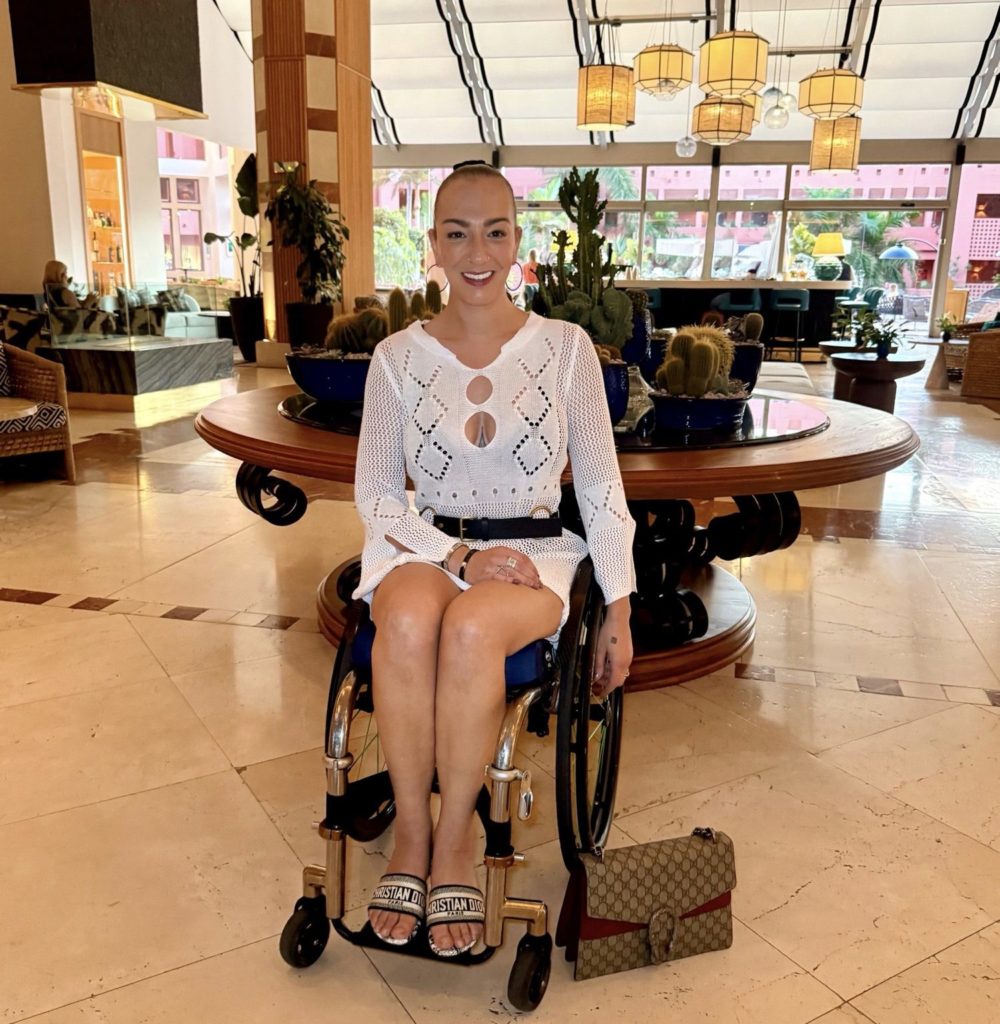 Luiz Faye seated in her wheelchair in the elegant lobby of The Ritz-Carlton Abama, wearing a white outfit, with decorative lanterns and a circular wooden display behind her.